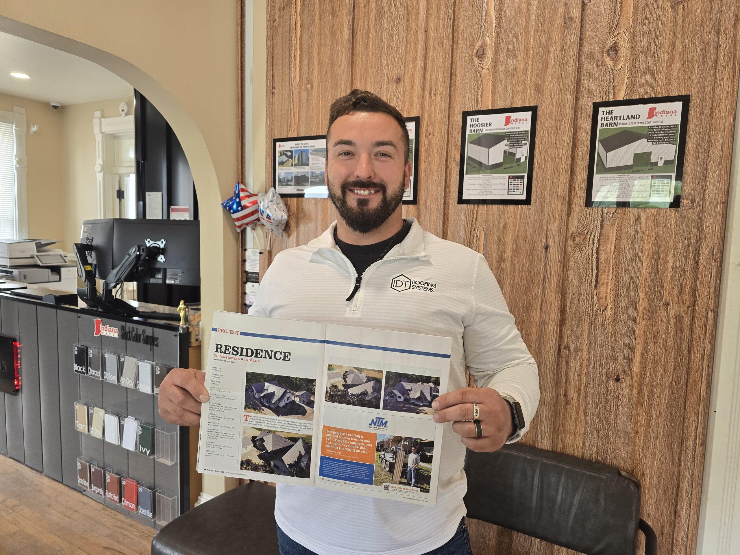 Cody Heller Showcases Featured Metal Roofing Project Cody Heller, owner of IDT Roofing Systems, proudly holding a Metal Roofing Magazine feature showcasing a recent residential project, standing inside Indiana Metal’s showroom with product displays and brochures visible in the background